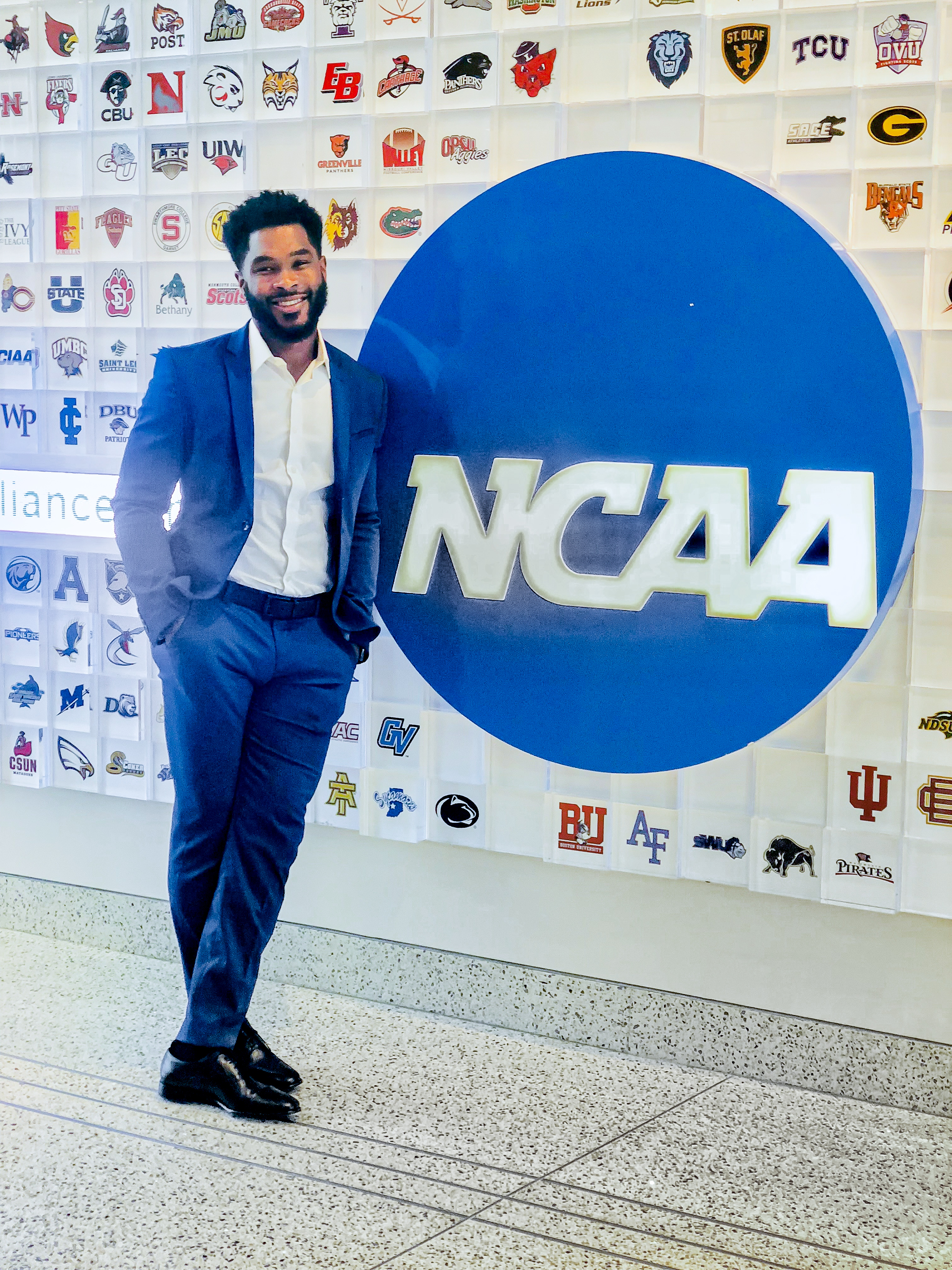 Brandon Johnson in a navy suit standing beside a large NCAA logo, in front of a wall of college athletics emblems
